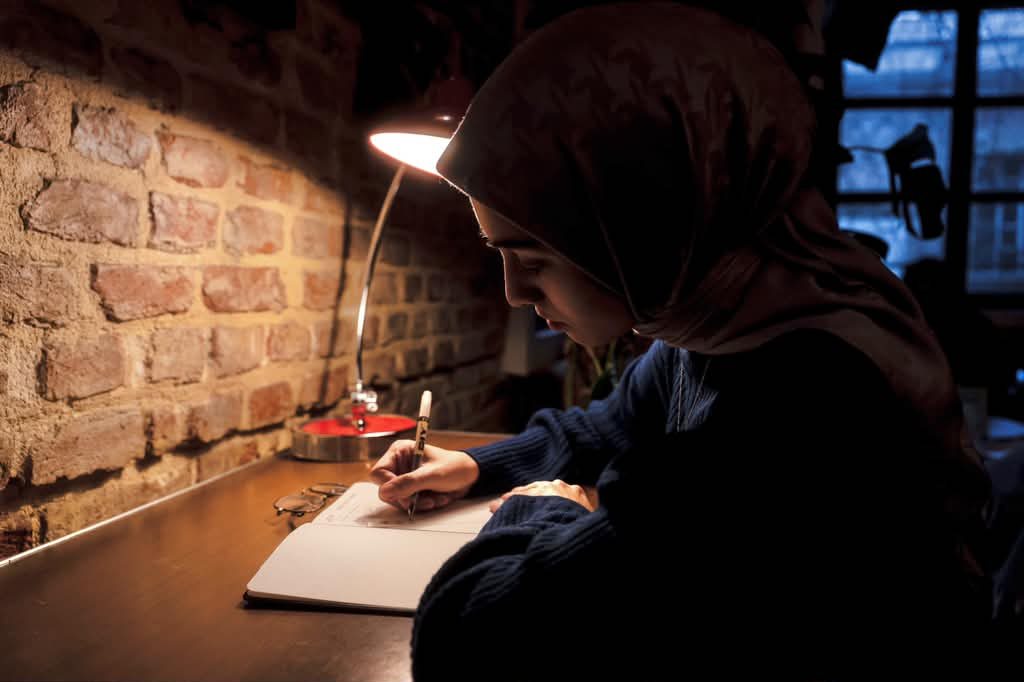 A young woman in a headscarf writing in a notebook by lamplight